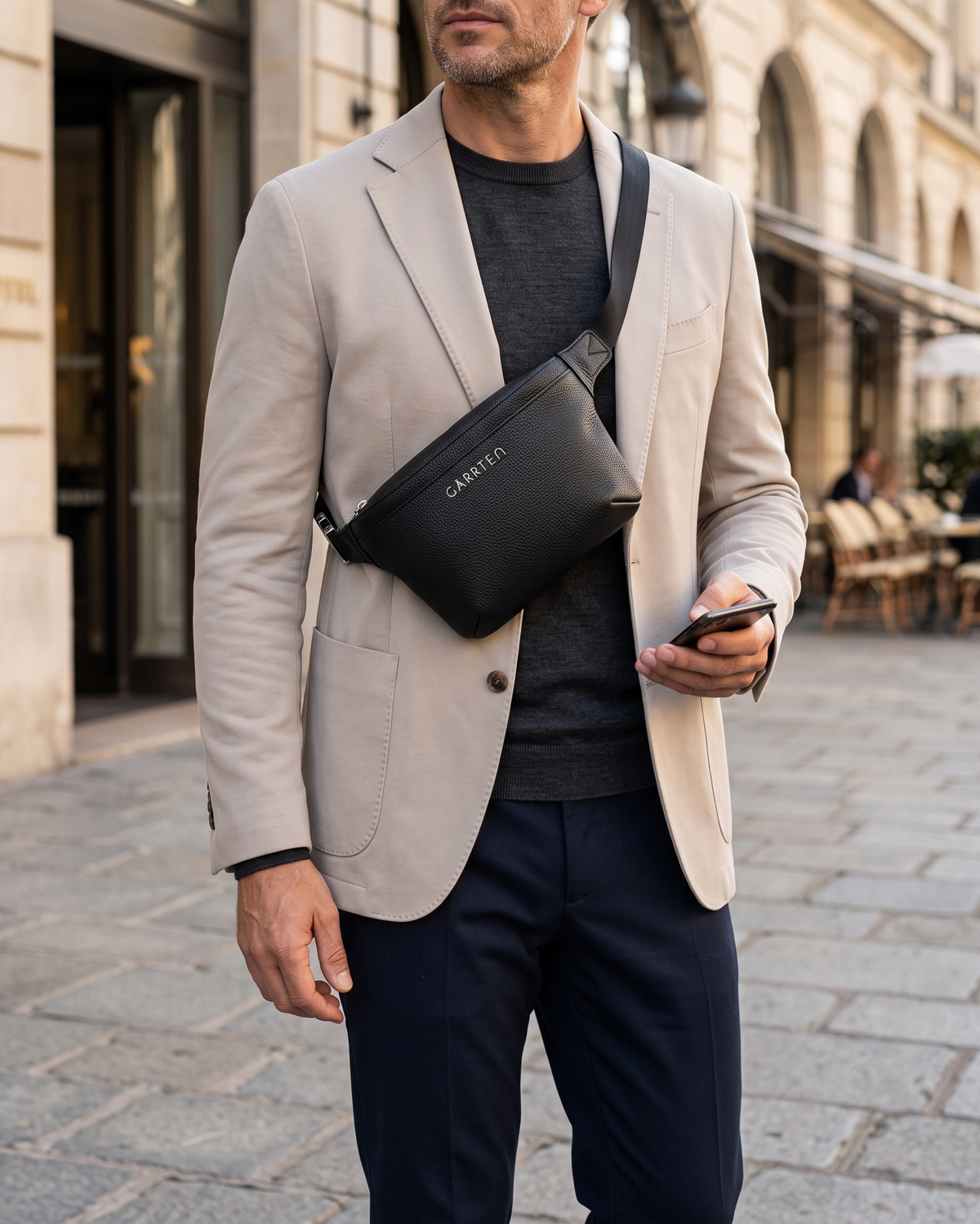 A man wearing a light beige blazer, dark gray t-shirt, and navy trousers carries a black leather crossbody bag with the brand name 'GARRTEN' in silver metal lettering, walking on a cobblestone street.